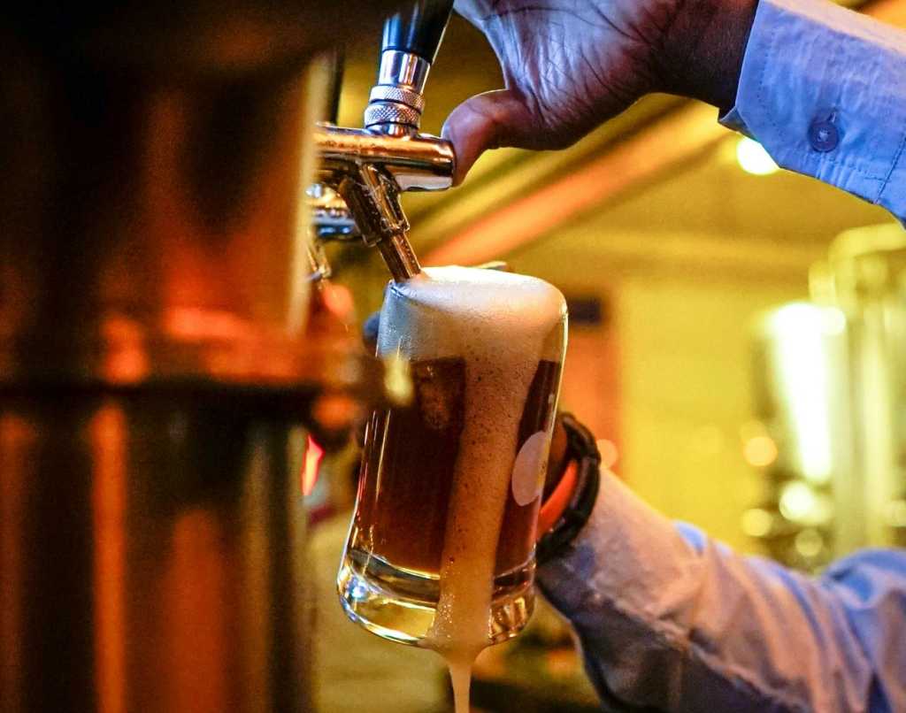 Hands filling a beer glass with drink from a beer tap. White foam leaks down one side of the glass.