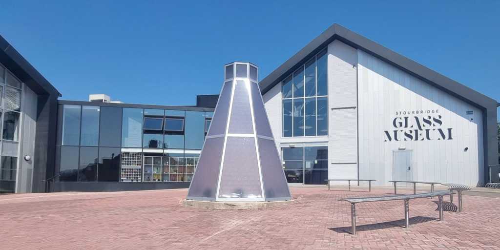 Modern white silver and glass buildings, with a red brick courtyard in the foreground and a silver-coloured cone sculpture in the centre