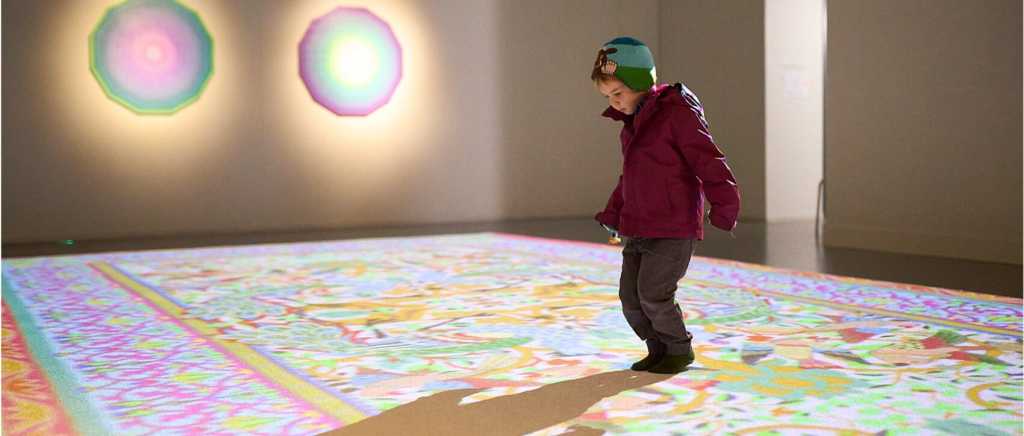 Child walking over a colourful rug on the floor of an art gallery, with two circular coloured lights on the wall behind